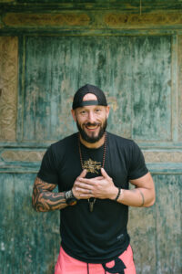 A cheerful, athletic man with a beard and tattoos posing warmly during a outdoor lifestyle portrait session.